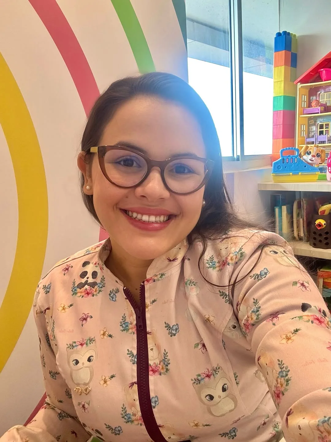 Yasmim sorrindo, de óculos e jaleco rosa estampado, na sala de atendimento com arco-íris na parede e brinquedos na estante