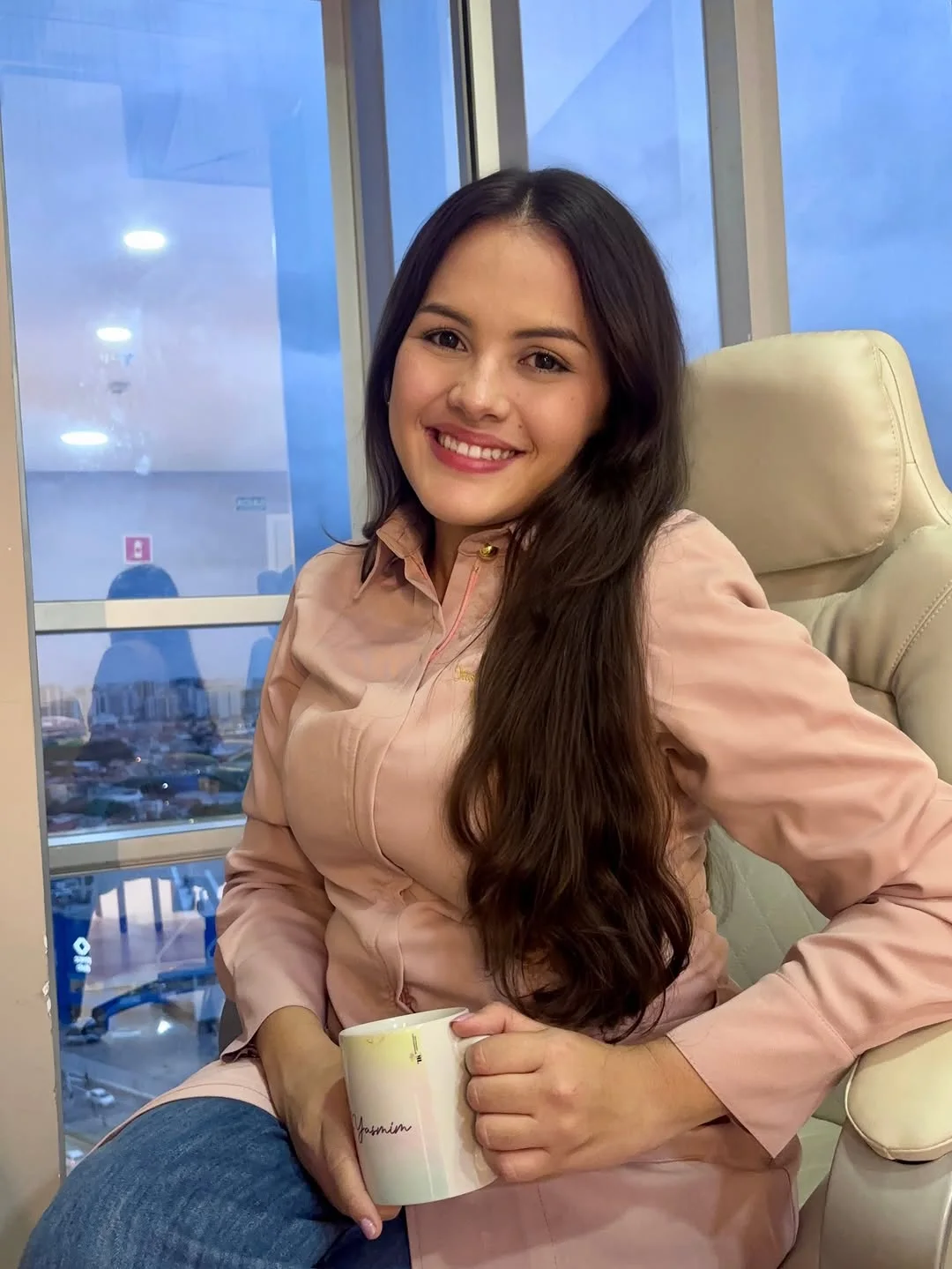 Yasmim Albuquerque sorrindo, sentada perto da janela, com uma caneca nas mãos