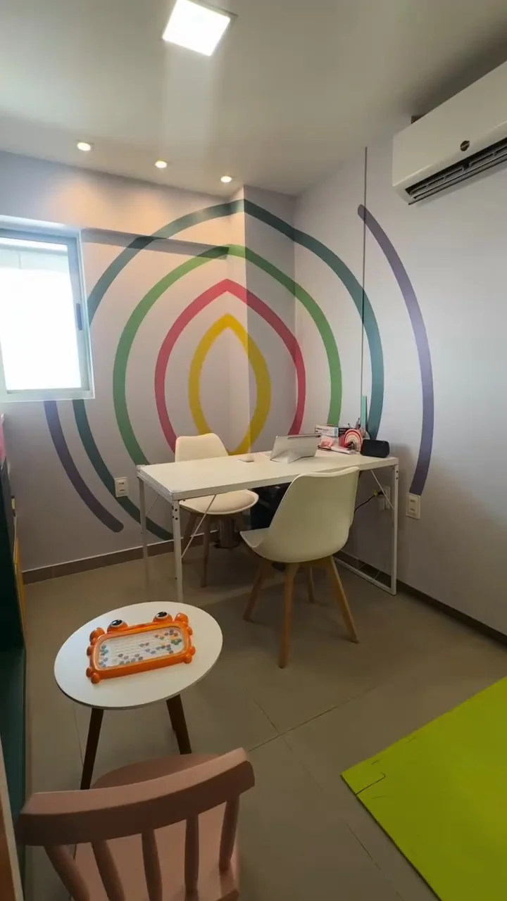 Sala de atendimento com mesa, cadeiras infantis, lousa mágica e arco-íris na parede