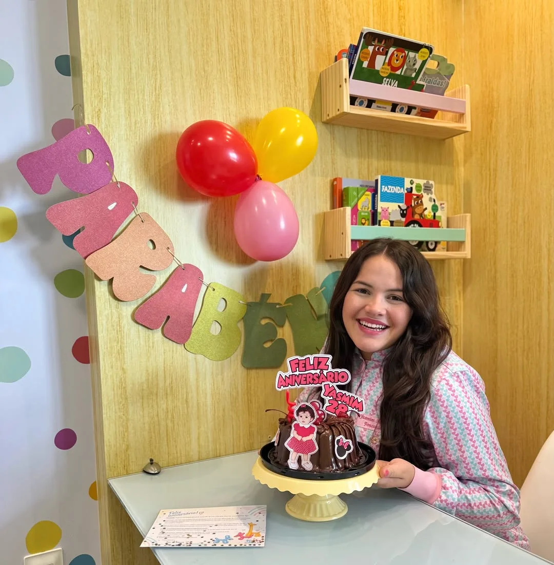 Yasmim sorrindo à mesa, com um bolo de aniversário, balões e um varal de letras na parede