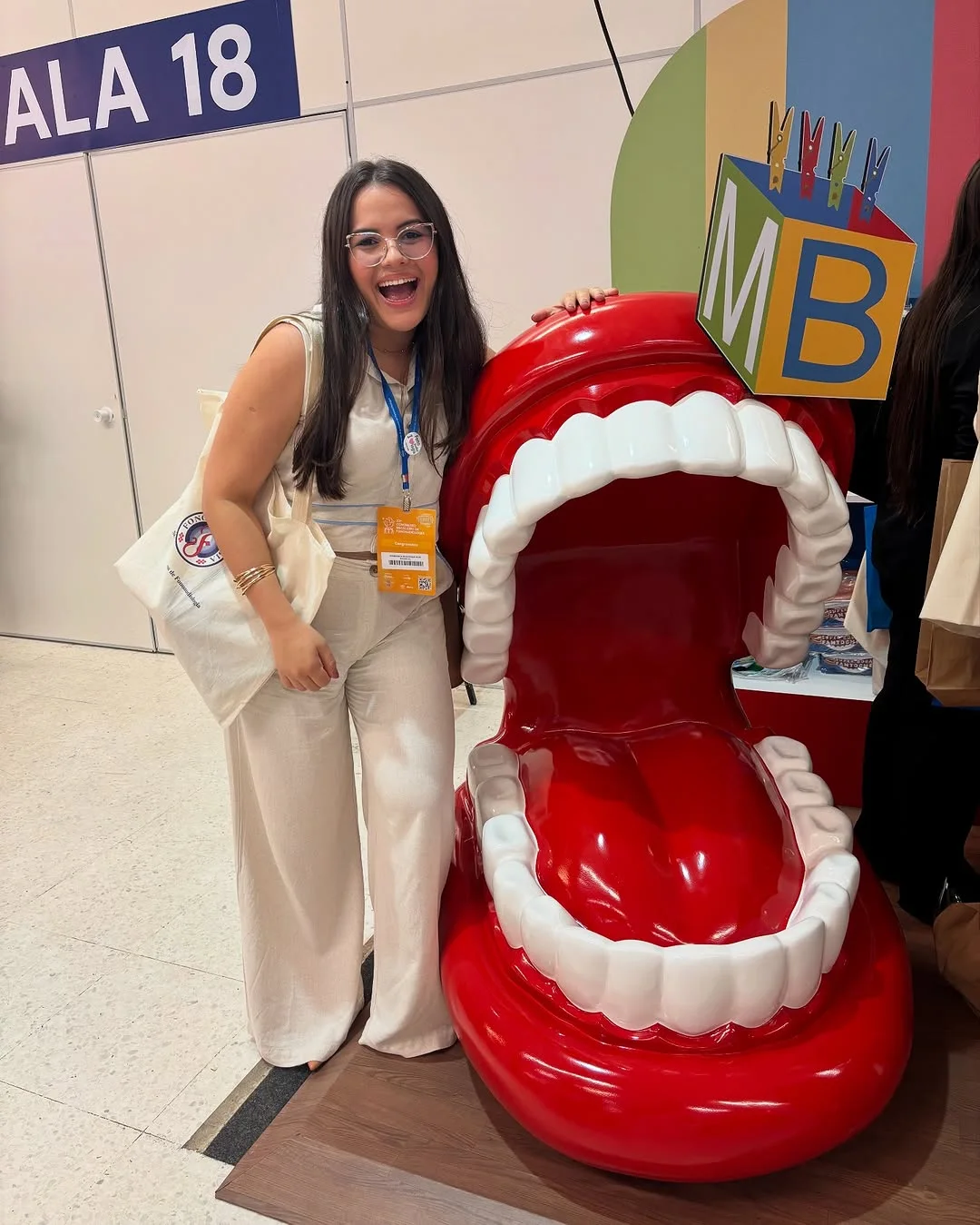 Yasmim rindo ao lado de uma boca gigante cenográfica no congresso de fonoaudiologia
