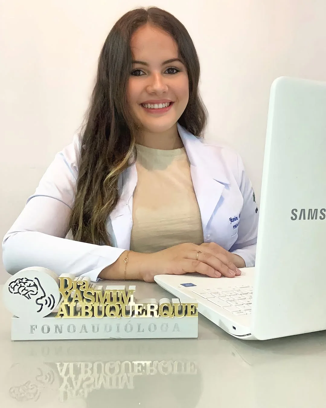 Yasmim em 2023, de jaleco branco, sorrindo à mesa com notebook e a placa com o nome dela